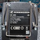 Černý vysílač s drátěným klipem. Na štítku je uveden nápis SENNHEISER, model SK 100 Bodypack Transmitter s označením G3. Jsou zde uvedeny údaje Prod.…
