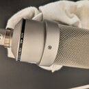 Stříbrný kondenzátorový mikrofon s kovovou síťovkou. Na těle je černý pruh s bílým nápisem SER. NO. 118273 a na spodní části je viditelné logo CE. Po…