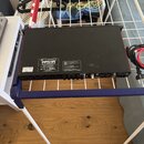 Černý elektronický přístroj s červenými bočními panely, umístěný na bílo-modrém sušáku. Na zadní straně jsou vidět různé konektory, přepínače a dvě i…