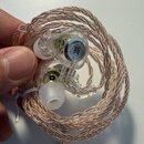 Sluchátka in-ear s průhledným plastovým tělem a měděně zbarveným pleteným kabelem. Na vnitřní straně jednoho sluchátka je viditelné černé kulaté logo…