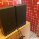 Dva černé hranaté reproduktory s látkovými kryty. Na spodní liště obou kusů je bílý nápis Schneider. Reproduktory jsou umístěny na světlém dřevěném n…