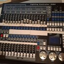 Černý světelný kontrolér s nápisem Lighting Controller 1024. Zařízení obsahuje množství posuvníků, tlačítek a tři velké stříbrné otočné ovladače v do…