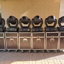 Šest černých moving head světel umístěných na šesti hnědých transportních kufrech s kovovými hranami a kolečky. Na spodní části světel jsou viditelné…