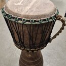 Djembe s tělem z tmavého dřeva a koženou blánou. Horní okraj je opleten zeleným šňůrkem a nástroj je napnutý černými provazníky. Na pravé straně je p…