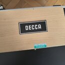 Fotografie zachycuje část černého transportního kufru s dřevěným panelem na víku. Na panelu je umístěna černá tabulky s bílým nápisem DECCA. V dolní…