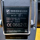 Záběr na zadní stranu šedého bezdrátového vysílače s kovovým klipem. Na černém štítku je uveden nápis SENNHEISER BODYPACK TRANSMITTER SK 100, FREQ. R…