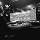 Černobílá fotografie zachycuje studio s osobou sedící u pracovního stolu. Na stole se nachází široký monitor s otevřeným softwarem pro tvorbu hudby,…