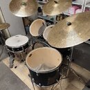 Černá bubnová sada s kovovým kováním a několika činely. Na jednom z činelů je viditelné logo Zildjian. Sada obsahuje snare, dva tom-tomy a basový bub…
