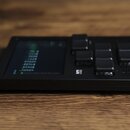 Černý kompaktní elektronický přístroj s displejem a několika ovládacími tlačítky. Na boku je viditelné logo MB a nápis SONG. Zařízení leží na dřevěné…