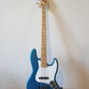 Modrá elektrická basová kytara s bílým pickguardem a světlou hnedou hmatníkem. Na hlavě nástroje je viditelné logo Fender a nápis MADE IN USA. Nástro…