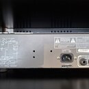 Zadní strana šedého elektronického zařízení s kovovým povrchem. Na horní části je schéma zapojení, ve spodní části se nachází vstup AC INPUT s nápise…