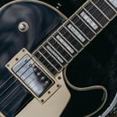 Detailní záběr na černou elektrickou kytaru v otevřeném kufru. Nástroj má hnědý hmatník s bílými blokovými vložkami a krémový pickguard. Na těle jsou…