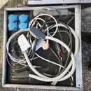 V hliníkovém transportním kufru se nachází svazek různých elektrických kabelů v bílé a černé barvě. V levé části jsou umístěny dvě modré zásuvky s kr…