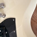 Detail korpusu elektrické kytary v krémové barvě s černým pickguardem. Jsou vidět kovové části mostu a struny. Na laku korpusu je v pravé části vidit…