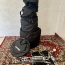 Fotografie zobrazuje sadu bubnového příslušenství a nástrojů. V horní části je stříbrný snare drum, pod ním jsou čtyři černé transportní vaky různých…
