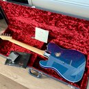 Modrá elektrická kytara typu Telecaster v otevřeném kufru s červeným plyšovým vnitřkem. Na kytaru je vidět výrazné odřapání laku na zadní straně korp…