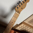 Hrabedlo elektrické kytary s přírodním dřevěným povrchem a hnedým hradlem. Na hlavě je bílý nápis Fender TELECASTER®. Vidět jsou šest ladících mechan…