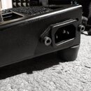Detailní záběr na zadní část černého elektronického zařízení s matným povrchem. Je zde vidět napájecí konektor typu IEC v plastovém rámečku, připevně…