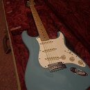 Světle modrá elektrická kytara typu Stratocaster s bílým pickguardem a javorovým hmatníkem. Nástroj leží v otevřeném kufru s červeným plyšovým vnitřk…