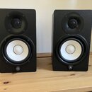 Dva černé studiové reproduktory s bílými membránami basových jednotek a černými výškovemi. V dolní části každého reproduktoru je umístěno kulaté logo…