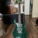 Zelená elektrická kytara s výraznou strukturou dřeva na těle a hnedým hmatníkem. Na hlavě je viditelné logo PRS. Kytara má dva humbuckery, fixační mo…