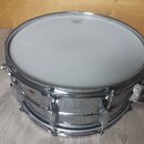 Kovový snare drum stříbrné barvy s bílým blátcem. Na obruči je viditelné logo znázornující korunu a nápis Ludwig. Povrch kovového kotle vykazuje znám…