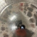 Detailní záběr na střed kovového činely zlatavé barvy s koncentrickým žebrováním. V uprostřed je vidět otvor se stříbrným šroubem. Povrch vykazuje zn…