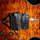 Detail elektrické kytary s oranžovým plamenovitým povrchem. Nástroj je vybaven dvěma černými humbuckery s nápisem ornpure a černým tremolo mostem. V…