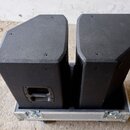 Dvě černé akustické reproboxy s texturovaným povrchem umístěné v transportním kufru z hliníku. Na přední straně levého reproduktoru je vidět nápis Tu…