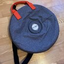 Kulatý šedý přepravní obal z textilního materiálu s oranžovými úchyty a černým popruhem. Uprostřed je umístěno kulaté logo s nápisem thomann a pod ní…