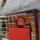Červený kovový box s bílými knoflíky na pravé straně. K boxu patří černý napájecí kabel s vidlicí a červený USB kabel. Vše je umístěno na bílé drátěn…