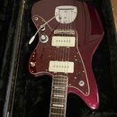 V černém kufru leží elektrická kytara v tmavě červené barvě s hnědo-oranžovým vzorovaným pickguardem. Nástroj má dva bílé humbuckery, vibrační systém…