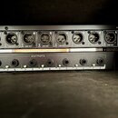 Černý rackový přístroj s řadou výstupů. Horní část obsahuje osm XLR konektorů označených čísly 1 až 8 a nápisy BAL OR UNBAL LINE OUT. Spodní část má…
