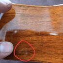 Detailní záběr na lakovaný dřevěný povrch hudebního nástroje v hnědé barvě. Červeným kroužkem je zvýrazněno oděrka laku v dolní části fotky. Na povrc…