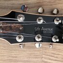 Detail hlavy kytary s černým povrchem a chromovanými ladičkami. Na hlavě je bílý nápis Godin a text 5th Avenue made in canada. V horní části jsou vid…