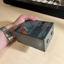 Šedý kovový efekt pedál v hranatém tvaru. Na horní straně je oranžový nápis ZOX a text X-BOX DI-BOX. Na boku jsou dva konektory s nápisy Input a Para…