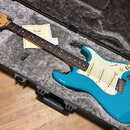 Modrá elektrická kytara typu Stratocaster s bílým pickguardem a hnědým hmatníkem, položená v šedém tvrdém kufru. Na hlavě kytary je viditelné logo Fe…