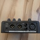 Zadávací panel černého elektronického zařízení s čtyřmi ovládacími knoflíky. Jsou zde viditelné konektory USB, dva XLR výstupy označené OUTPUT a THRU…