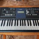 Černý digitální klavír značky YAMAHA s bílými a černými klávesami. Na ovládacím panelu jsou viditelné nápisy LESSON, STYLE a SONG spolu s různými tla…