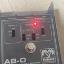 Šedý kovový přístroj s nápisem AB-O BALANCED LINE OUTPUT SWITCHER a logem Palmer Musical Instruments. Na horní straně jsou označení OUTPUT A, OUTPUT…
