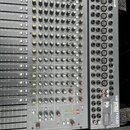 Šedý míchací pult s velkým množstvím posuvníků a otočných knoflíků v různých barvách. Na pravé straně je vidět panel s konektory a nápisy SPIRIT FOLI…