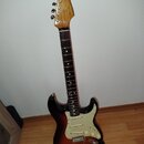 Elektrická kytara s tłem v barvě sunburst a bílým pickguardem. Na hlavě hředu je vidět nápis "Fender" a pod ním menší, nečitelný text. Kytara má hněd…