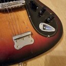 Detail těla elektrické basové kytary s červeným gradientem do černé. Nástroj má dřevěný povrch, černý pickguard s dvěma potenciometry a kovový most.…