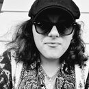 Na černobílé fotografii je zobrazen muž s dlouhými vlnitými vlasy, který má na sobě kšiltovku a sluneční brýle s nápisem Ray-Ban. Má na sobě vzorovan…