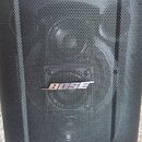 Černý cylindrický reproduktor s kovovou mřížkou. Uprostřed je umístěno stříbrné logo BOSE. Povrch mřížky vykazuje v pravé části viditelné zmačkání a…