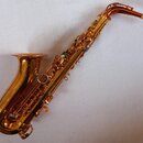 Zlatý saxofon s nablýskaným povrchem. Na zvonu je vidět rytiný ornament, ale žádný čitelný nápis ani značka. Nástroj je vybaven mouthpiecem s nasazen…
