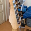 Fotografie zachycuje zadní stranu hlavy kytary z přírodního světlého dřeva. Na hlavě jsou osazeny šest chromovaných ladících mechanismů. Povrch dřeva…