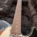 Elektrická kytara s černým bodies a hnědým hmatníkem, položená v černém plišovém kufru. Na těle je vidět bílý pickguard s výraznými odřapáním a opotř…