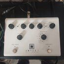 Bílý kytarový pedál s nápisem AMPED 1 a pod ním menším textem DEPT. 10 | 100W AMP PEDAL. Nad nápisem je logo ve tvaru hvězdy v černém čtverci. Zaříze…