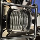 Černý kabel namotaný na kulatém černém cívce, která je umístěna v černém kovovém rámu. Na povrchu kabelu jsou v několika místech viditelné bílé nápis…